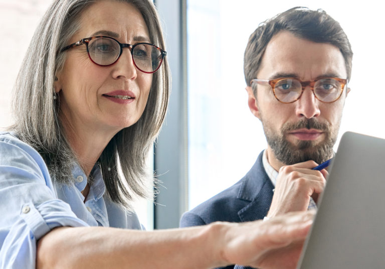 Mature older ceo businesswoman mentor in glasses negotiating growth business plan with diverse executive managers at boardroom meeting table using laptop. Multicultural team work together in office.